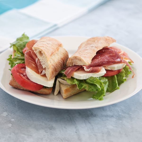 Two halves of a sandwich with prosciutto, mozzarella, tomato, and lettuce on a white plate.