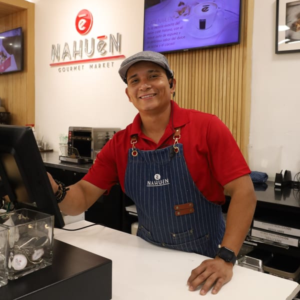 A smiling NAHUEN Gourmet Market employee in a red shirt and striped apron stands behind a counter.
