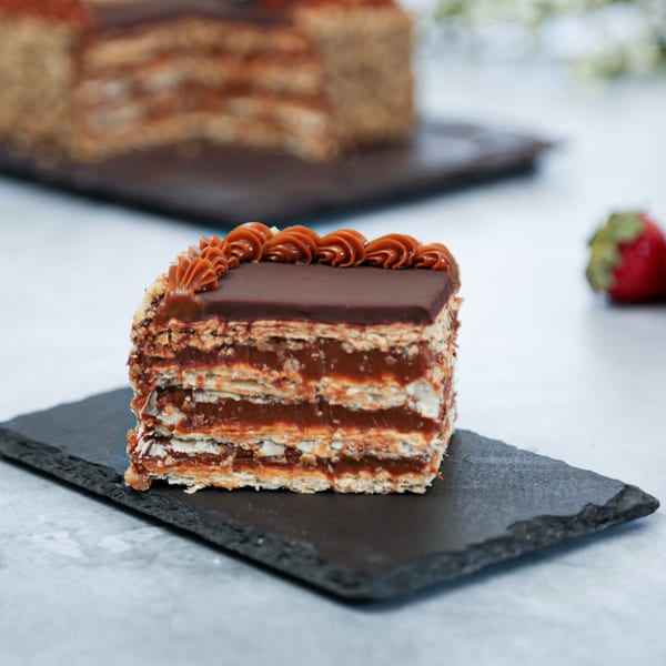 A slice of Argentinian dulce de leche cake with chocolate topping and a whole cake in the background.