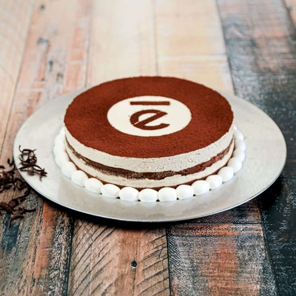 Round tiramisu cake with a white logo on top, surrounded by white meringue kisses on a silver platter.