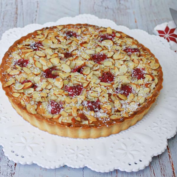 Round raspberry and almond tart on a white scalloped plate, with a red and white napkin.