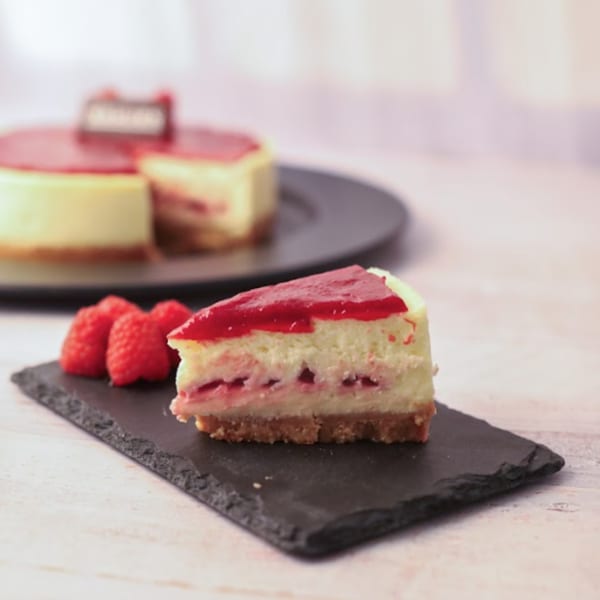 A slice of raspberry cheesecake with a whole cheesecake in the background, on a dark slate board.