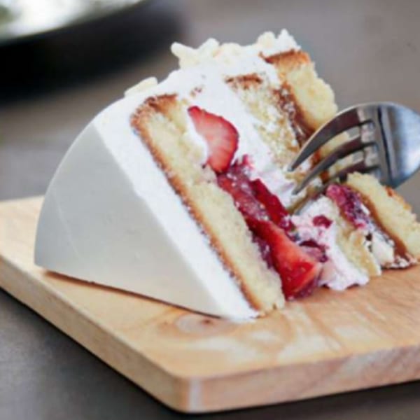 A fork digging into a slice of strawberry shortcake with white frosting on a wooden board.