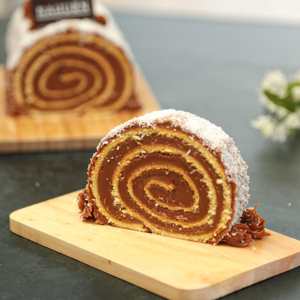 A slice of dulce de leche roll cake with coconut flakes on a wooden board.
