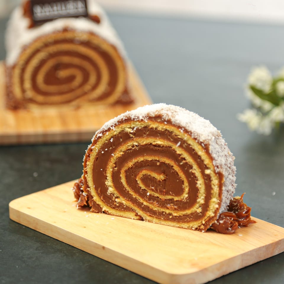 A slice of dulce de leche roll cake with coconut flakes on a wooden board.