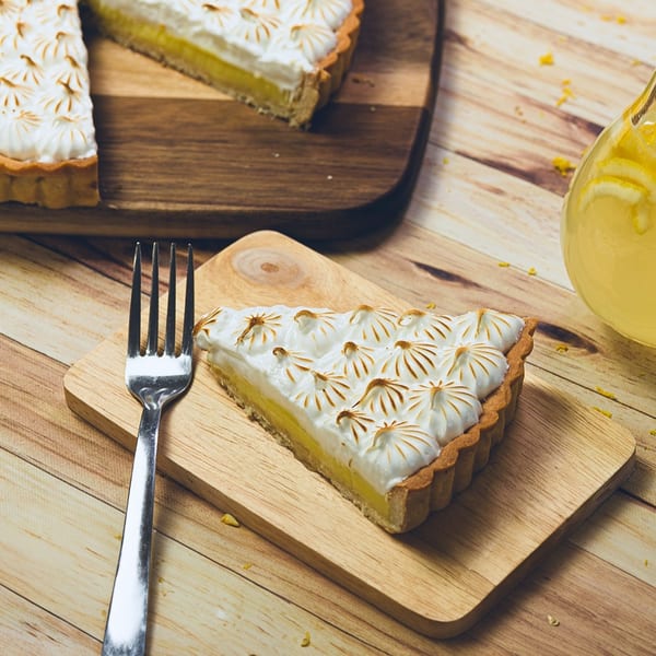 A slice of lemon meringue pie with toasted peaks on a wooden board next to a fork.