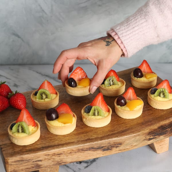 Hand placing a fruit tartlet with strawberries, kiwi, and blueberries on a wooden board.