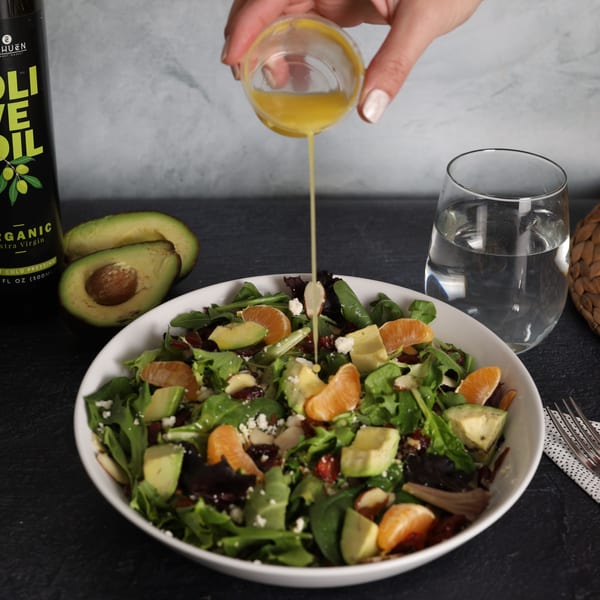 A hand pours dressing onto a fresh salad with avocado, mandarins, and feta, next to NAHUEN olive oil.