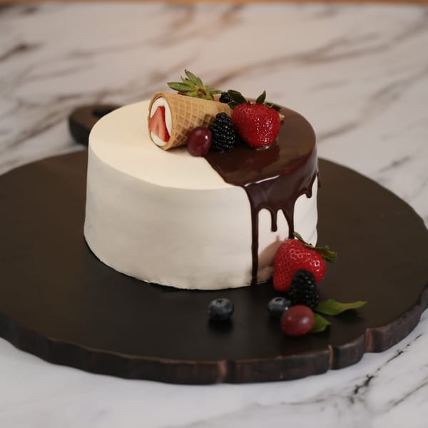 White frosted cake with chocolate drip, topped with strawberries, blackberries, grapes, and a waffle cone.