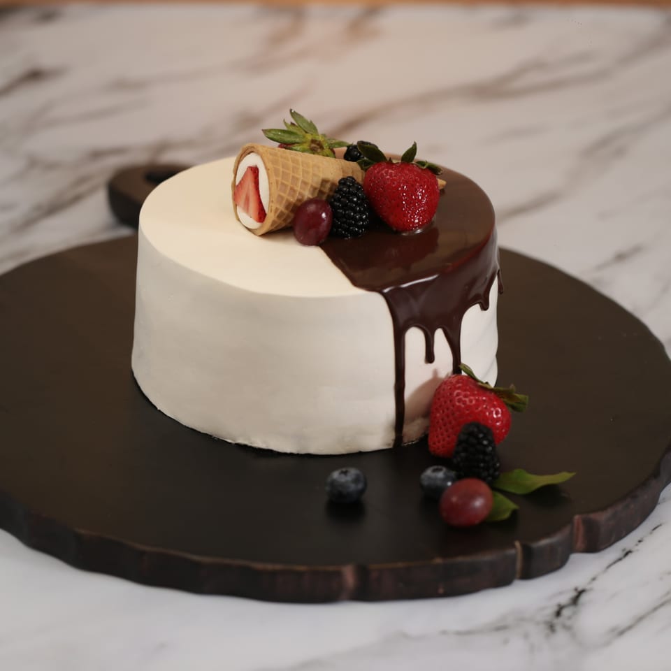 White frosted cake with chocolate drip, topped with strawberries, blackberries, grapes, and a waffle cone.