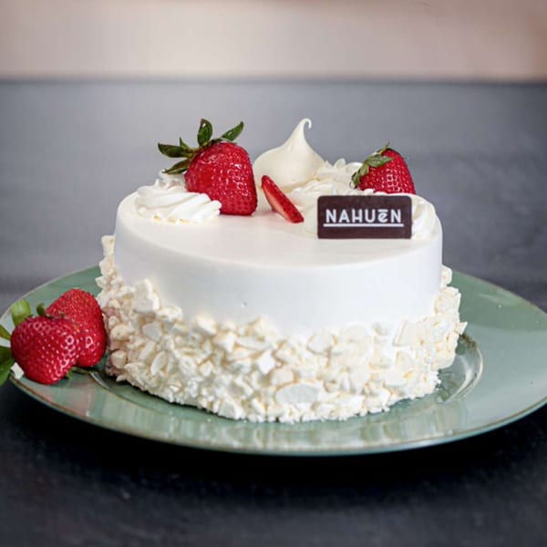 White Nahuen cake with meringue crumbles, topped with whipped cream, strawberries, and a chocolate plaque.