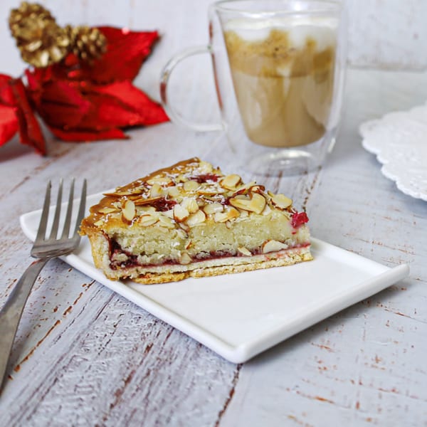Slice of Argentinian cake with raspberry filling and almond topping, with a latte and festive decor.