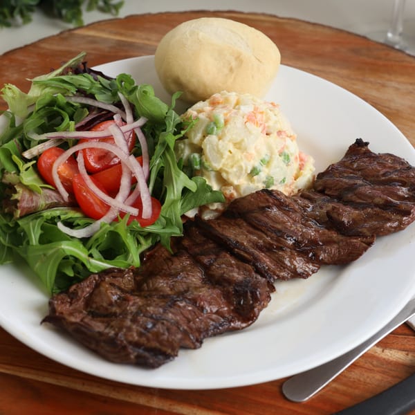 Grilled Argentinian steak with a side salad, potato salad, and a bread roll on a white plate.