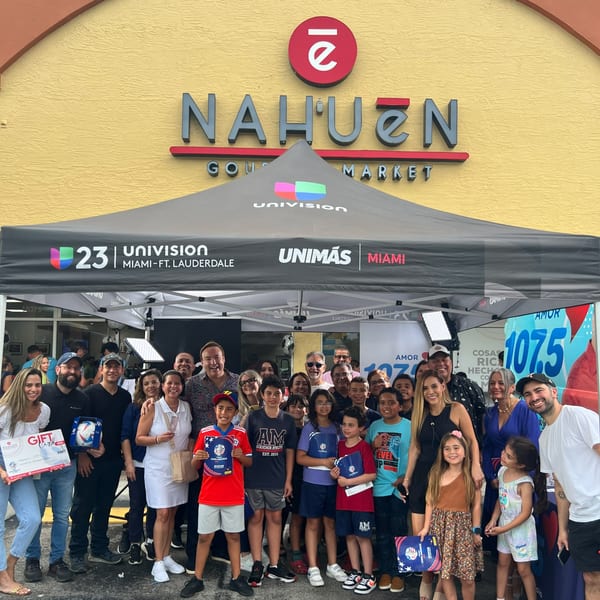 A large group of adults and children pose under a Univision tent in front of Nahuen Gourmet Market.
