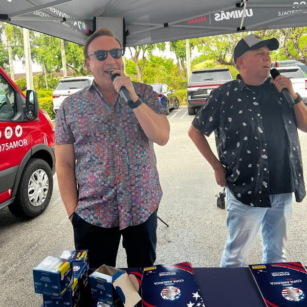 Two men speaking into microphones under a tent, with promotional materials on a table.