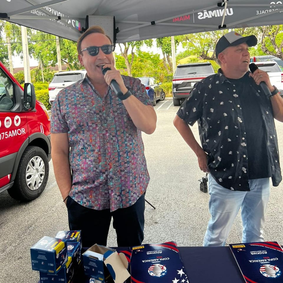 Two men speaking into microphones under a tent, with promotional materials on a table.