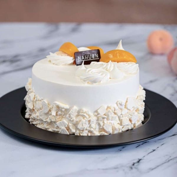 A white NAHUEN cake with meringue pieces, whipped cream, and peach slices on a black plate.