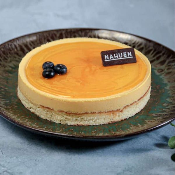 Round Nahuen cake with orange topping, blueberries, and a chocolate tag on a dark plate.