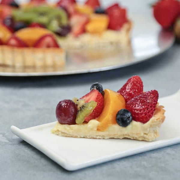A slice of fruit tart with strawberries, blueberries, kiwi, and peaches on a white plate.